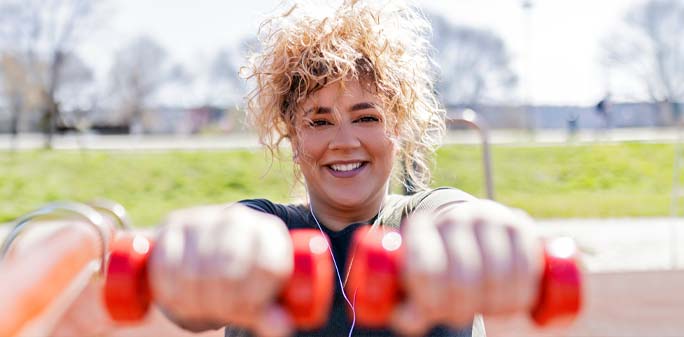 A woman smiles, exercising with two dumbbells outside